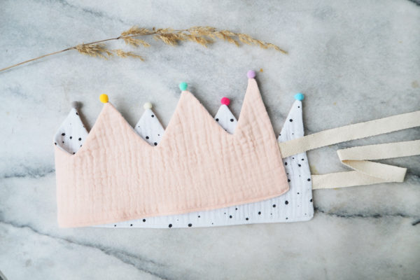 Eine Musselin Stoffkrone für Kinder, in pastellrosa und Innenseite weiß mit schwarzen punkten und Bindeschnur, auf einem wunderschönen Marmortisch. An den Kronspitzen sind winzige, bunte Pompon Details